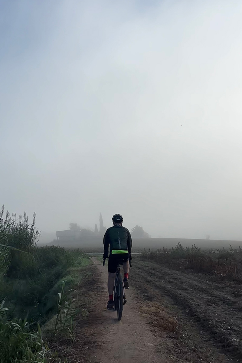 ciclista de gravel en un sendero estrecho en un día de niebla y una masia de un pueblo en el fondo